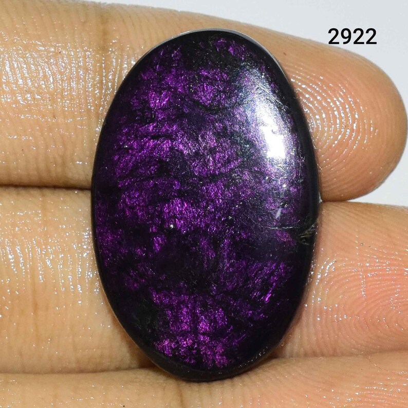 Puede incluir: Una piedra preciosa ovalada cabuj&oacute;n de color p&uacute;rpura oscuro con brillo iridiscente. La piedra tiene un fondo negro con motas p&uacute;rpuras y plateadas.