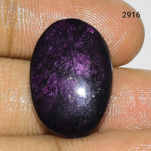 Puede incluir: Una piedra preciosa ovalada cabuj&oacute;n de color p&uacute;rpura oscuro con un acabado brillante e iridiscente. La piedra tiene una base negra con motas p&uacute;rpuras y plateadas.