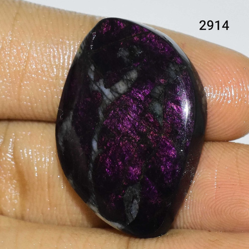 Puede incluir: Un cabuj&oacute;n de piedra preciosa de color morado oscuro y negro con una superficie lisa y pulida. La piedra tiene un brillo iridiscente &uacute;nico y un patr&oacute;n de remolino.