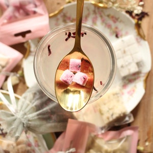 May include: A gold spoon holds three pink sugar cubes with dried flower petals in a glass jar. The sugar cubes are in a clear glass jar with a white background.