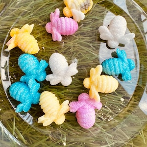 May include: A collection of ten sugar cookie bee shapes in various colors, including yellow, blue, pink, white, and a black and yellow bee. The cookies are decorated with sugar crystals and are arranged on a clear plate.