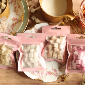 May include: Four small pink pouches with clear windows, each containing different colored sugar cubes. The pouches are on a white plate with a floral pattern. There is a cup of tea, a cup of milk, and a bowl of sugar cubes in the background.