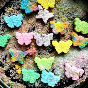 May include: A collection of colorful butterfly-shaped bath bombs in a black bowl. The bath bombs are covered in glitter and are arranged in a random pattern.