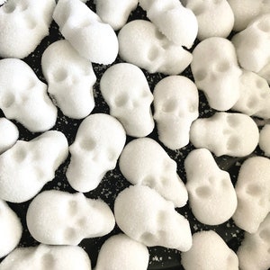 May include: A close-up shot of numerous white, skull-shaped candies, each covered in a layer of granulated sugar. The candies are scattered across a dark surface, creating a stark contrast. The image evokes a Halloween or Day of the Dead theme.