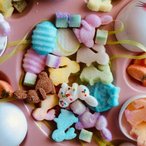May include: A variety of pastel-colored sugar candies shaped like bunnies, eggs, and other Easter-themed shapes. The candies are arranged on a pink tray with white egg-shaped containers.