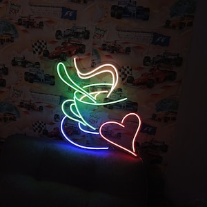 Könnte beinhalten: Ein Neon-Schild in Form einer Kaffeetasse mit aufsteigendem Dampf. Die Tasse ist grün, die Untertasse ist blau und der Dampf ist weiß. Ein rotes Herz befindet sich neben der Tasse.