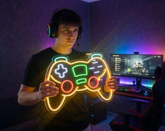 May include: A neon-lit gaming controller in yellow, green, red, and white is held by a person wearing a black t-shirt and headphones. The controller's outline glows brightly. A computer setup is visible in the background, suggesting a gaming theme.
