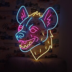 May include: A neon sign in the shape of a hyena's head, with a blue outline and pink and yellow accents. The sign is mounted on a wall with a racing car wallpaper.