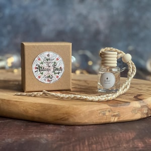 May include: A clear glass air freshener bottle with a wooden cap and a jute cord, beside a small brown box. The box features a round sticker with the text "Flicker The Addams Family". The items are on a wooden board.
