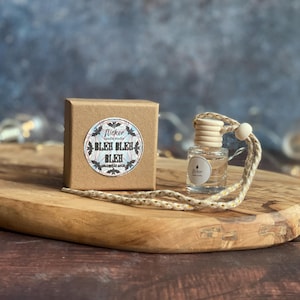 May include: A small glass bottle with a wooden top and a rope, alongside a brown box. The box features a circular sticker with the words "BLEH BLEH BLEH" and a bat design. The items are displayed on a wooden board, with a blurred background.