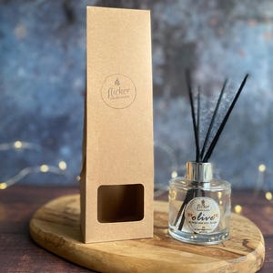 Könnte beinhalten: Eine klare Glas-Diffusorflasche mit schwarzen Schilfrohren und einem silbernen Deckel, neben einer braunen Pappschachtel mit einem Logo. Die Flasche hat ein Etikett mit der Aufschrift "olive". Die Artikel befinden sich auf einem Holztablett.