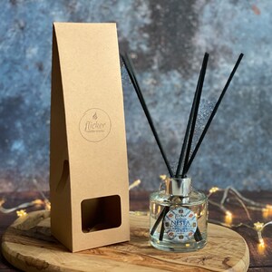 Könnte beinhalten: Ein klarer Glasdiffusor mit schwarzen Schilfrohren und einem blau-weißen Etikett mit der Aufschrift "Flicker Candle Co. Nesta - Sea Salt & Sage - 100ml". Der Diffusor steht auf einer Holzoberfläche mit einem braunen Karton dahinter.