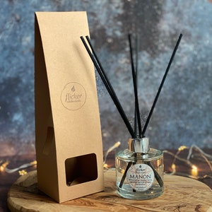 May include: A clear glass diffuser bottle with black reeds and a silver cap, labeled "Manon" with "Grapefruit" below. A brown cardboard box with the "flicker candle studio" logo is to the left. The diffuser sits on a wooden surface.