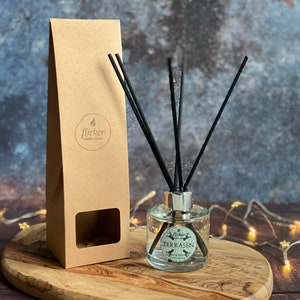 Puede incluir: Un difusor de vidrio transparente con cañas negras y una etiqueta blanca que dice "flicker candle studio" y "TERRASEN" en verde. El difusor está sobre una superficie de madera con una caja de papel marrón detrás.