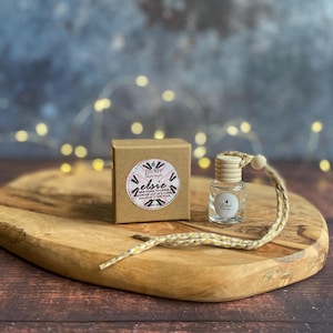 Könnte beinhalten: Eine kleine, klare Glasflasche mit einem Holzverschluss und einer geflochtenen Schnur, neben einer kleinen braunen Schachtel. Die Schachtel hat ein rundes Etikett mit Text. Die Artikel befinden sich auf einem ovalen Holzbrett.