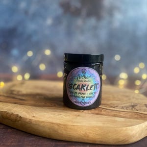 May include: A black jar candle with a black lid, labeled "Scarlett" with a colorful circular sticker. The candle sits on a wooden cutting board. The background is blurred with bokeh lights.