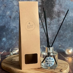 May include: A clear glass diffuser bottle with black reeds and a silver collar, next to a brown cardboard box. The bottle has a label that reads "The Haunted Mansion". The box has a window and a logo that says "flicker candle studio".