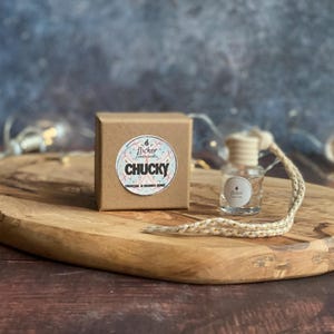 May include: A brown box with a sticker reading "flicker candle studio" and "CHUCKY" is on a wooden board. A small glass bottle with a wooden cap and a string is next to the box. The background is blurred with fairy lights.