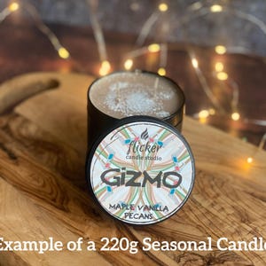 May include: A black candle tin with a lid featuring the word "Gizmo" and the scent "Maple Vanilla Pecans." The candle sits on a wooden cutting board. The text "Example of a 220g Seasonal Candle" is at the bottom.