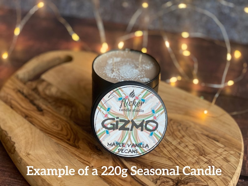 May include: A black candle tin with a lid featuring the word "Gizmo" and the scent "Maple Vanilla Pecans." The candle sits on a wooden cutting board. The text "Example of a 220g Seasonal Candle" is at the bottom.