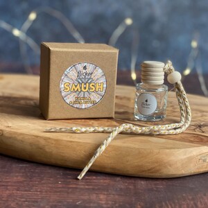 May include: A clear glass bottle with a wooden cap and a decorative string, next to a brown cardboard box. The box has a circular label with the word "SMUSH" and "Vanilla & Shea Butter" printed on it. The items are on a wooden tray.