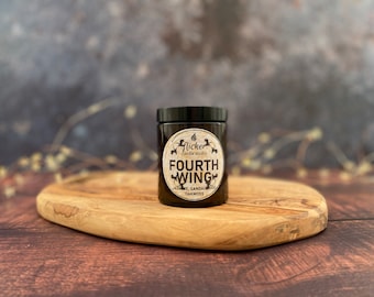 May include: A dark amber glass candle jar with a black lid. The label reads "flicker candle studio" and "FOURTH WING" in a white font. The scent description is "jasmine, sandalwood, oakmoss".