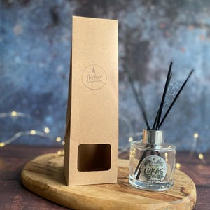Könnte beinhalten: Ein klarer Glas-Diffusor für ätherische Öle mit schwarzen Stäbchen und einem silbernen Deckel, neben einer braunen Pappschachtel. Der Diffusor hat ein Etikett mit der Aufschrift "Lukas". Die Box hat ein Fenster und ein Logo "flicker candle studio".