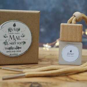 May include: A brown cardboard box with a white sticker that says "flicker candle studio, MAL I'LL MEET YOU AT THE MEADOW HIMALAYAN CEDAR & JASMINE". Next to the box is a small glass bottle with a wooden top and a beige cord. The bottle has a white sticker that says "flicker candle studio".