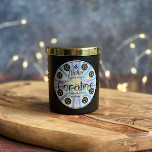 May include: A black candle with a gold rim, labeled "Coraline" with "Strawberry & Chocolate Cake" scent. The candle is on a wooden cutting board. The label features a circular design with a button motif and the text "flicker candle studio."