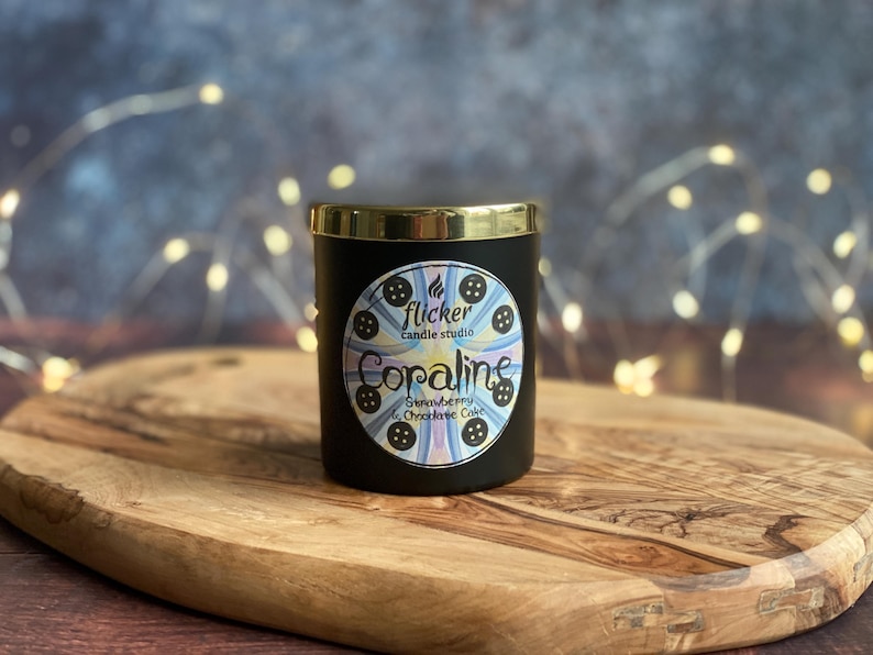 May include: A black candle with a gold rim, labeled "Coraline" with "Strawberry & Chocolate Cake" scent. The candle is on a wooden cutting board. The label features a circular design with a button motif and the text "flicker candle studio."
