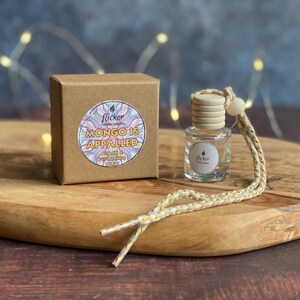 May include: A small glass bottle with a wooden stopper and a decorative string, beside a brown cardboard box. The box features a sticker with the text "MONGO IS APPALLED" and "flicker". The items are on a wooden board.