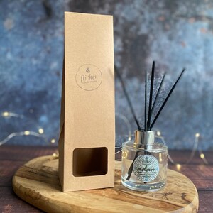 Könnte beinhalten: Ein klarer Glas-Aromatherapie-Diffusor mit schwarzen Schilfrohren und einem silbernen Kragen steht auf einem Holzbrett. Links befindet sich eine hohe, braune Pappschachtel mit einem Fenster und einem Logo. Der Diffusor hat ein Etikett mit der Aufschrift "Adam".