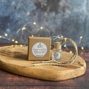 Puede incluir: Una pequeña botella de vidrio transparente con una tapa de madera y un cordón tejido, junto a una pequeña caja marrón. Ambos artículos tienen el logotipo "flicker candle studio". La caja tiene una etiqueta con texto. Los artículos están sobre una tabla de cortar de madera.