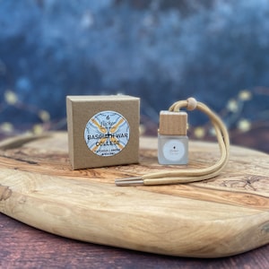 Puede incluir: Una caja de cartón marrón con una etiqueta blanca que dice "Flicker Candle Studio" y "Basgiath War College Patchouli Amber & Cocoa". Una pequeña botella de vidrio con una tapa de madera y un cordón beige se encuentra sobre una tabla de cortar de madera.