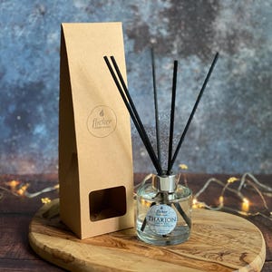 Puede incluir: Un difusor de vidrio transparente con varillas negras y un collar plateado, junto a una caja de papel marrón. La botella tiene una etiqueta que dice "Tharion". La caja tiene el logo "flicker". Sobre una superficie de madera.