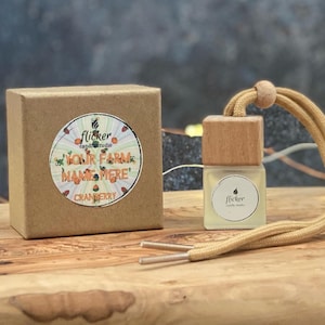 Può includere: Una scatola di cartone marrone con un adesivo bianco che dice "Flicker candle studio" e "Your Farm Name Here! Cranberry". Un piccolo deodorante per auto in legno con uno spago beige è accanto alla scatola. Il deodorante ha una bottiglia di vetro trasparente con un adesivo bianco che dice "Flicker candle studio".