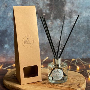 Könnte beinhalten: Eine klare Glasflasche mit silbernem Deckel und schwarzen Diffusorstäbchen. Die Flasche ist mit einer klaren Flüssigkeit gefüllt und trägt ein Etikett mit der Aufschrift "Gwyn I am the Rock..." Die Flasche steht auf einer Holzoberfläche, neben ihr befindet sich eine braune Papiertüte.