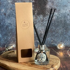 Könnte beinhalten: Eine klare Glas-Diffusorflasche mit schwarzen Schilfrohren und dem Text "Lets Split Up". Die Flasche steht auf einem Holztablett neben einer braunen Pappschachtel mit einem Logo. Dekorative Lichter befinden sich im Hintergrund.