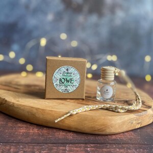 Könnte beinhalten: Eine kleine, klare Glasflasche mit Holzverschluss und dekorativer Schnur, neben einer kleinen braunen Schachtel. Beide Artikel haben einen runden Aufkleber mit dem Text "flicker candle studio" und "IOWE".