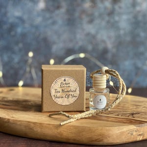 Op de afbeelding: Een klein bruin doosje met een wit label waarop staat "Flicker Candle Co. Ten Hundred Years Of You". Een heldere glazen fles met een bruine dop en een ketting van houten kralen hangt aan het doosje.