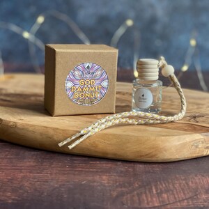 May include: A small, clear glass bottle with a wooden cap and a decorative string, next to a brown box. The box has a circular sticker with the text "GOD DAMMIT DONUT". The items are on a wooden tray.