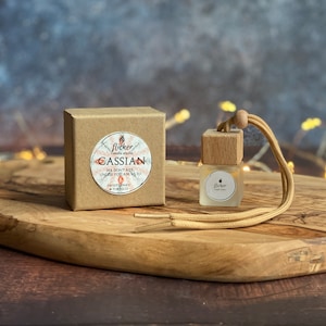 Könnte beinhalten: Eine braune Pappbox mit einem weißen Etikett, auf dem "Flicker Candle Studio Cassian We don't ask, unless you ask us to Sweet Honey & Tobacco" steht, und eine kleine braune Holzflasche mit einem durchsichtigen Glasfläschchen, das einen Autolufterfrischer mit einer braunen Schnur enthält.
