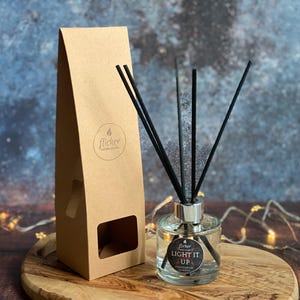 May include: A clear glass diffuser bottle with black reeds and a brown paper box. The bottle has a silver cap and a black label that reads "Flicker Light It Up Sandalwood & Cedarwood Diffuser".