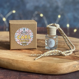 May include: A small, clear glass bottle with a wooden cap and a woven cord, next to a brown cardboard box. The box has a circular label with the text "CARL BOOM! Tobacco & Whisky". The bottle and box are on a wooden surface.