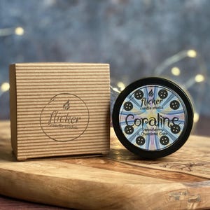May include: A candle studio product with a brown cardboard box and a black tin candle. The candle tin has the name "Coraline" and the scent "Strawberry & Chocolate Cake". The box and candle are on a wooden surface.