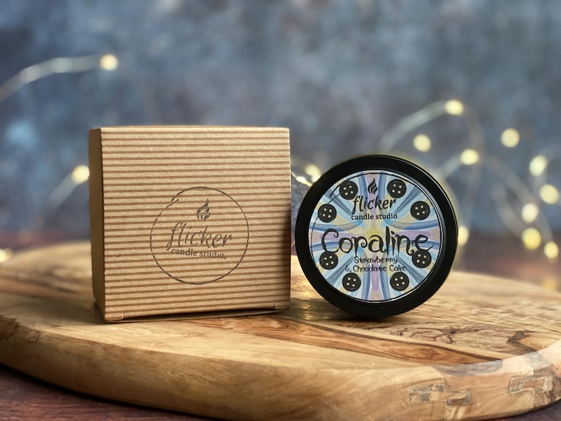 May include: A candle studio product with a brown cardboard box and a black tin candle. The candle tin has the name "Coraline" and the scent "Strawberry & Chocolate Cake". The box and candle are on a wooden surface.