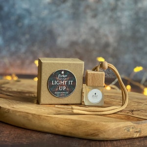 Könnte beinhalten: Eine kleine Holzkiste mit einem braunen Papieretikett, auf dem "Flicker Candle Diffuser Light It Up Passionfruit Martini" steht. Eine kleine Glasflasche mit einem Holzdeckel und einer braunen Schnur hängt an der Kiste.