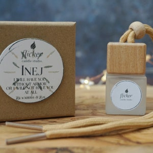 May include: A brown cardboard box with a white circle sticker that says "flicker candle studio" and "INEJ I WILL HAVE YOU WITHOUT ARMOR OR I WILL NOT HAVE YOU AT ALL BY LEIGH BARDUGO". A small glass bottle with a wooden top and a white circle sticker that says "flicker candle studio" is attached to a tan cord.