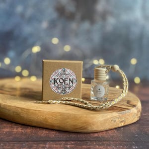Könnte beinhalten: Eine kleine Glasflasche mit Holzverschluss und geflochtener Kordel neben einer kleinen braunen Schachtel mit dem Wort "KOEN". Die Artikel befinden sich auf einem Holztablett. Der Hintergrund ist mit Bokeh-Lichtern verschwommen.