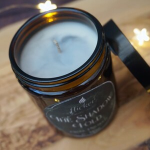 May include: A brown glass jar candle with a black label that reads "Flicker & Co. The Shadow Cloud Soy Wax Candle". The candle is lit and the wax is melting.